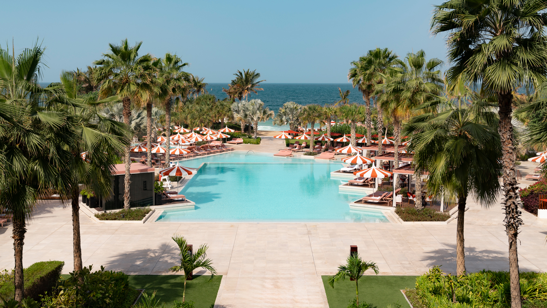 Scenic view of a luxurious beachfront resort pool surrounded by palm trees and orange-striped umbrellas, overlooking the sea under a clear blue sky.